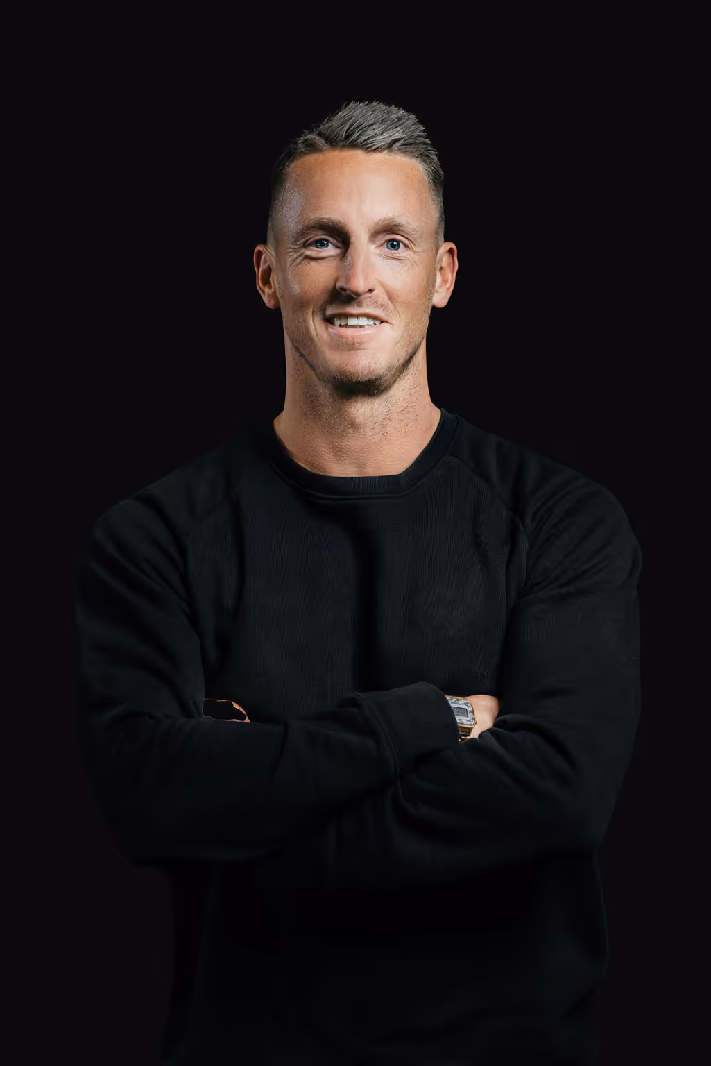 Smiling man with short gray hair wearing a black sweater and silver watch standing with arms crossed against a black background.