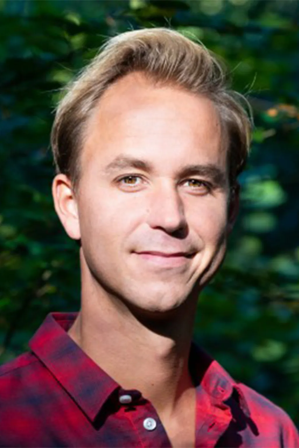 Close-up of a smiling man with blonde hair wearing a red and black checkered shirt against a green leafy background.