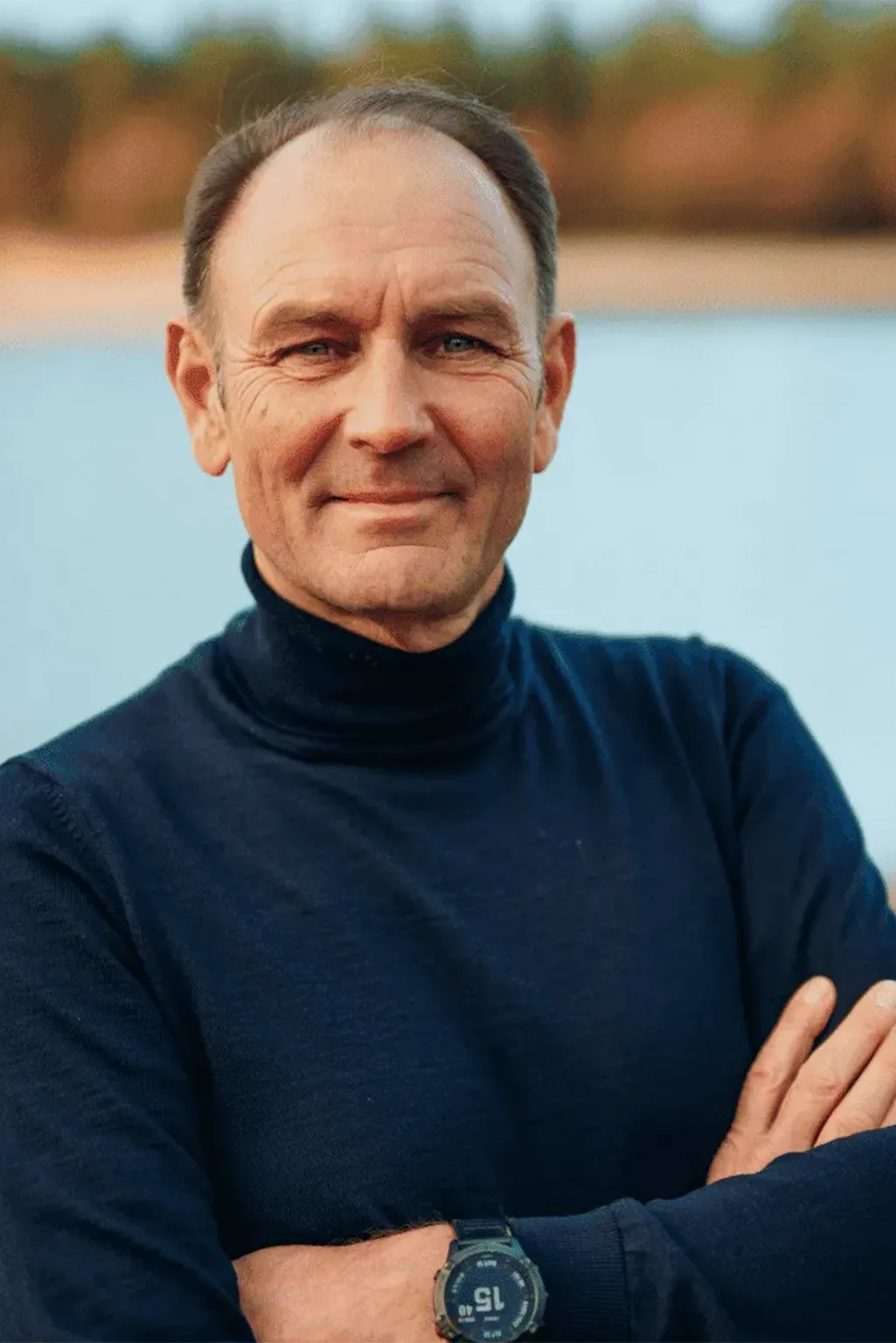 Close-up of a smiling man with blonde hair wearing a red and black checkered shirt against a green leafy background.