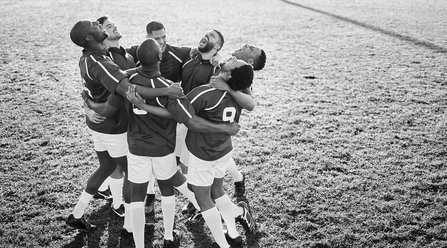 A group of male soccer players in uniform embracing in a circle on a grassy field, looking upwards and shouting joyfully.